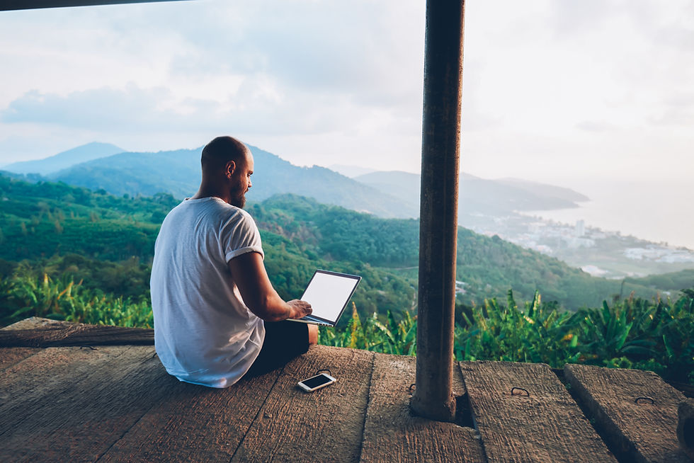 Man in a white shirt working on a laptop on a wooden deck overlooking lush green hills and a distant cityscape; phone nearby. Serene mood.